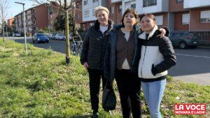 Tre donne, commercianti del quartiere Sant'Agabio, sorridono abbracciate stando in piedi su un'area verde a bordo strada. Indossano abbigliamento casual invernale. Alle loro spalle si nota una strada asfaltata con automobili in transito, un ciclista di passaggio, una fila di alberi spogli e dei condomini residenziali con facciate di colore rosso mattone e bianco.