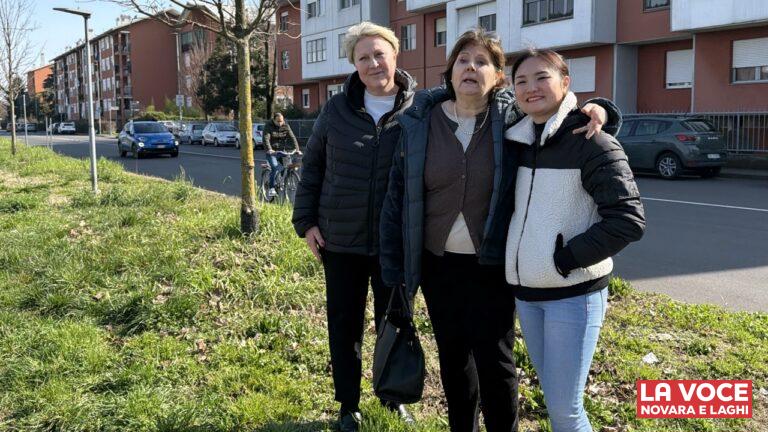 Tre donne, commercianti del quartiere Sant'Agabio, sorridono abbracciate stando in piedi su un'area verde a bordo strada. Indossano abbigliamento casual invernale. Alle loro spalle si nota una strada asfaltata con automobili in transito, un ciclista di passaggio, una fila di alberi spogli e dei condomini residenziali con facciate di colore rosso mattone e bianco.